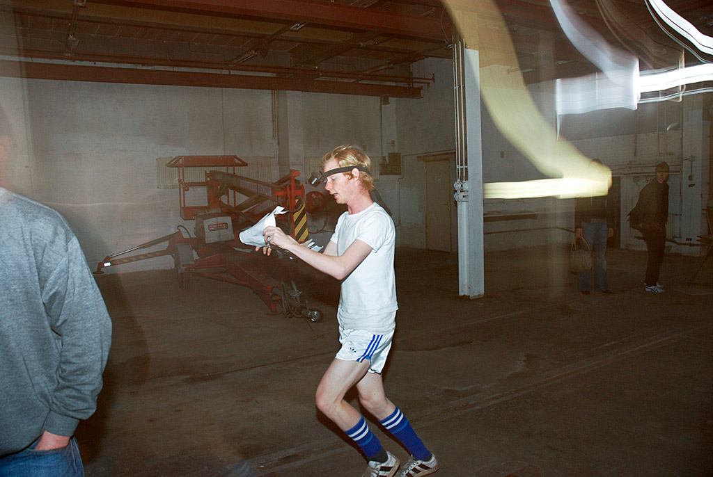 You see a young man running through an industrial space. Dressed in sportswear, the most striking garment being his Adidas shorts. While running he appears to be reading a text from a large piece of paper which he is holding in his hands and which is lit by a light mounted on his forehead. The look in his eyes is somewhat strained. The scene is framed by onlookers from all sides, who’s static posture contrasts the runner’s motion.
