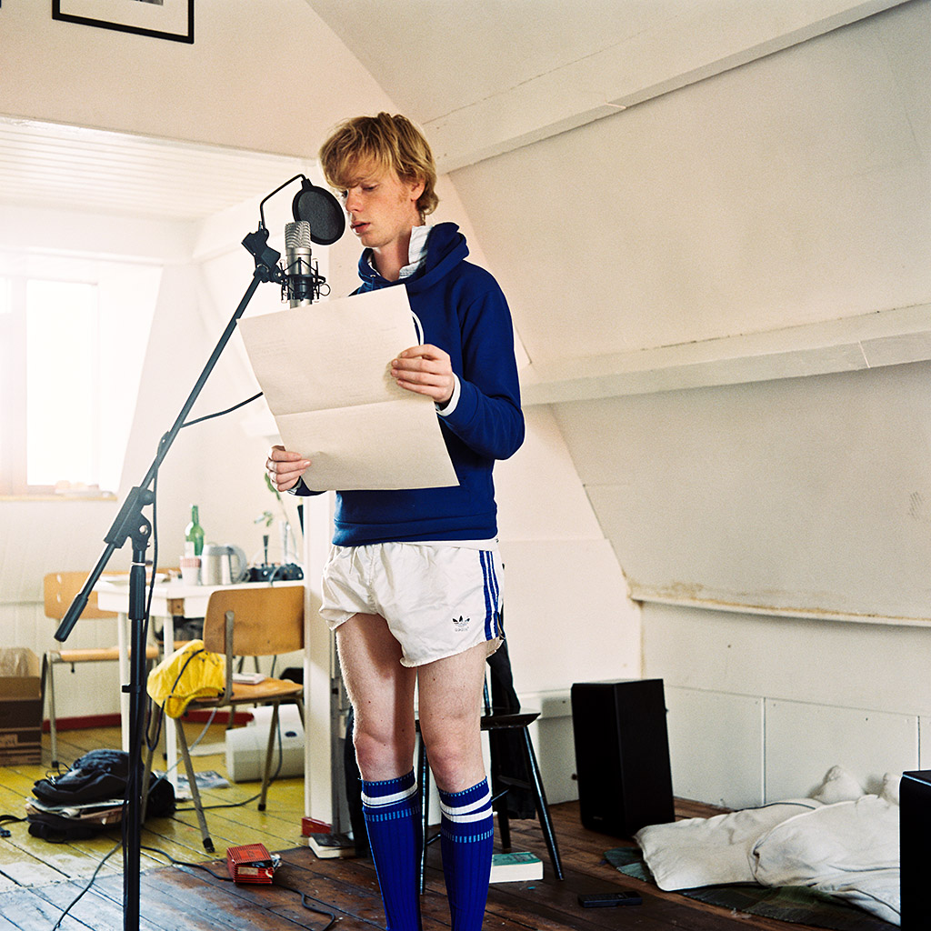 You see a young man standing in the living room of a small appartment. He appears to be speaking into a broadcasting microphone that is mounted on a microphone stand. He is holding a large sheet of yellowish paper from which he appears to be reading. He’s wearing a blue hooded sweater on top of a pair of glossy white running shorts. A striking detail are the young man’s legs. It is obvious he never wears shorts, as his legs are pallid and goosy, especially his upper legs. One leg of the shorts is pent-up.