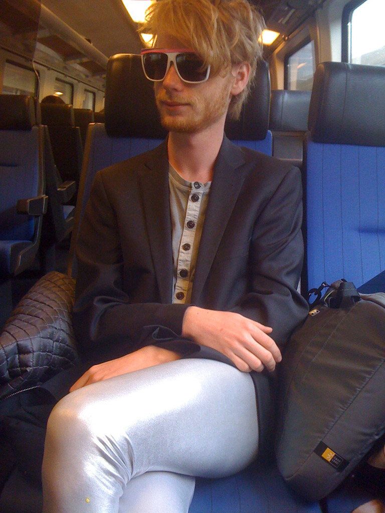You see a young man in a train wearing white and purple sunglasses. He wears a jacket and silver tights and has a content air.