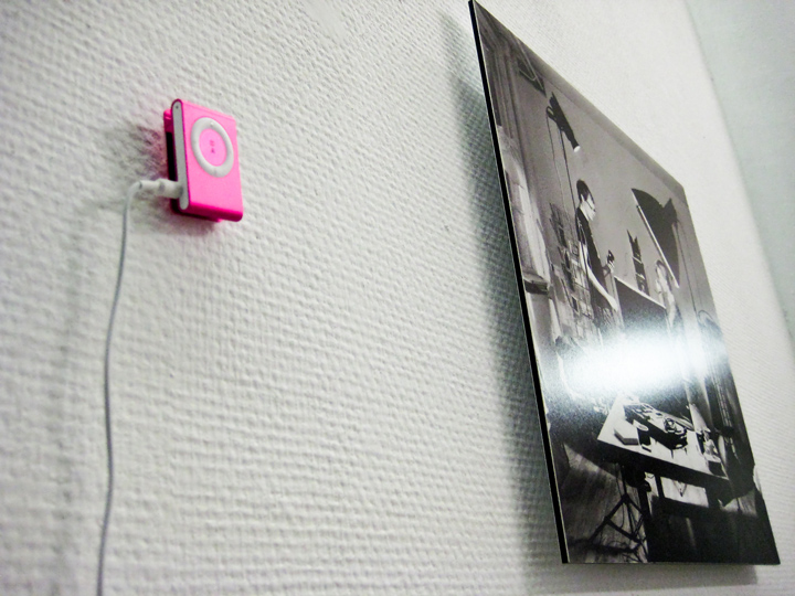 You see a close up of a gallery wall. The depth of field is shallow. There's a small pink iPod in the foreground, and a photographic black and white print in the background which is mounted on a thick plate-like material. Neither of these is in focus however; the plane of focus lies exactly on the edge of the photographic print.