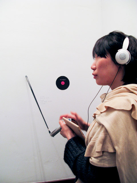 You see a Korean woman in her late-twenties, pensively listening to sound on white headphones. She is holding a portable cd player in her hands. In the background, on the wall she is facing, you see a handwritten text and a stylised representation of a vinyl record, with a pink label.