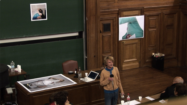 You see an old-fashioned university auditorium covered in wooden panels. A man wearing colourful garments smiles at the onlookers. He holds a microphone in his hands. The blackboard, the wall, and a desk in front are covered by large pictures. The pictures depict men who somehow hide their face.