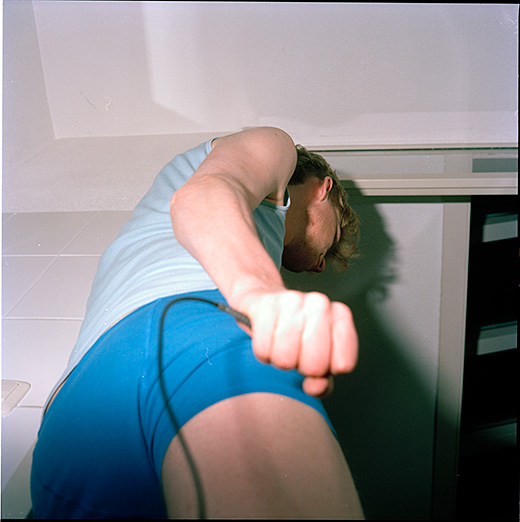 You see a young man standing in what appears to be a bathroom. He is wearing a blue tank top and blue shorts. You see him from beneath, in profile, starting from the thighs. You can not see his eyes. His right hand holds a remote release firmly. The flash of the camera has overlit his hand and produced a large shadow to his right.
