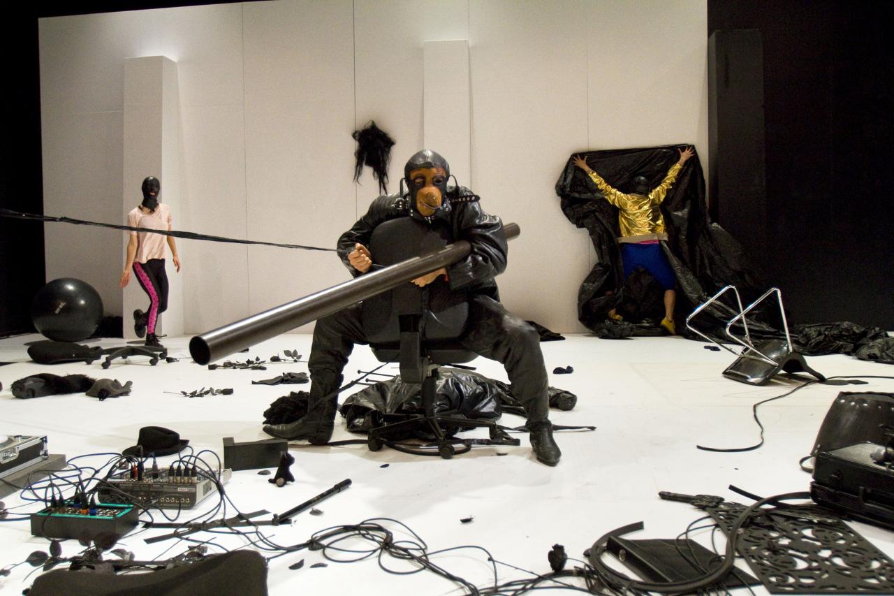 You see a stage littered with black props. Different kinds of plastic, a chair overturned, a mixing desk… The ballet floor is white as is the wall that is erected in the back. In the background two characters are wearing black latex masks. One of them is walking towards the centre.