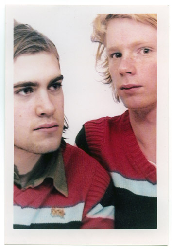 You see a polaroid picture of two young men wearing identical pink sweaters. It's a posed picture, the background is white, there are no identifiable surroundings.