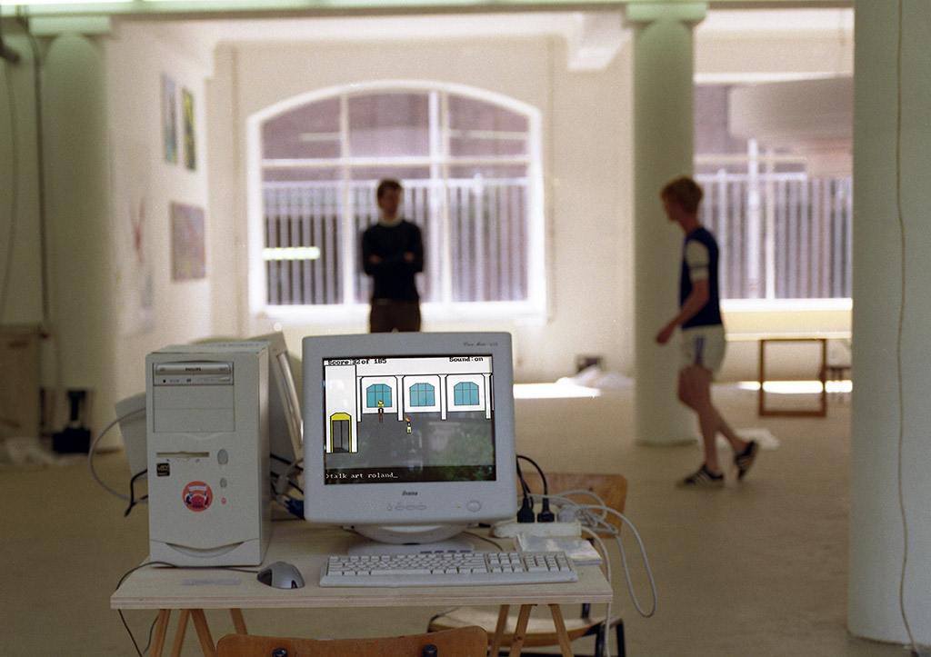 You see a picture of an industrial space, apparently used as an exhibition site for visual art. A computer is visible in the foreground, upon which you can make out the same screenshot you just saw. Behind the computer, out of focus, you see a person in shorts leaning towards a tall person who stands up straight.