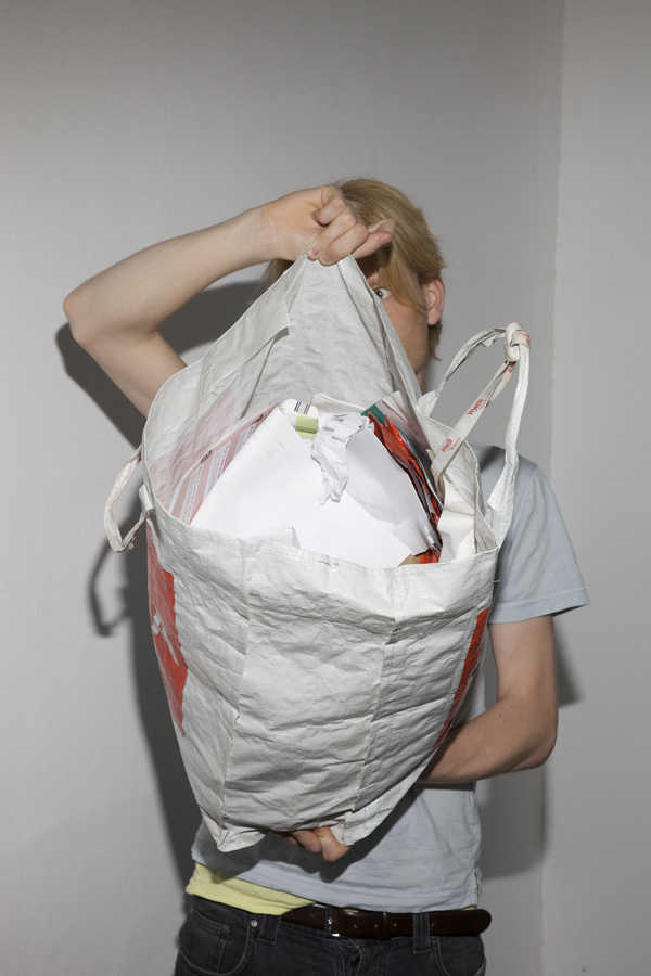 You see a young man holding a large plastic bag in front of his face and torso. The bag is filled with paper waste.