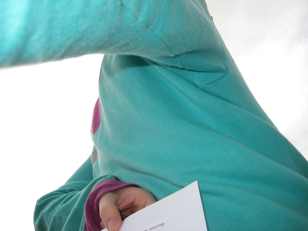 You see the back of a man wearing a colourful hooded sweater. He holds a small piece of paper clenched in his left hand, which is pressed to his back.